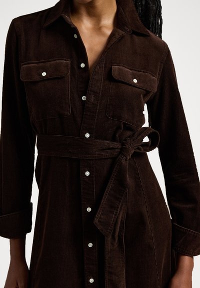Brun kordfløjlskjole med lange ærmer, krave, to brystlommer, knaplukning foran og matchende bælte. Har tekstureret stof og hvide knapper.