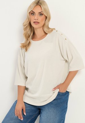Blondynka ubrana w luźną kremową prążkowaną bluzkę z guzikami na jednym ramieniu i niebieskie dżinsy, stoi z jedną ręką w kieszeni.