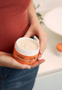 weDo/ Restore & Shine Mask in een ronde, oranje tub met een witte deksel. Romige, dikke consistentie zichtbaar van binnen. Inhoud: 150 ml.