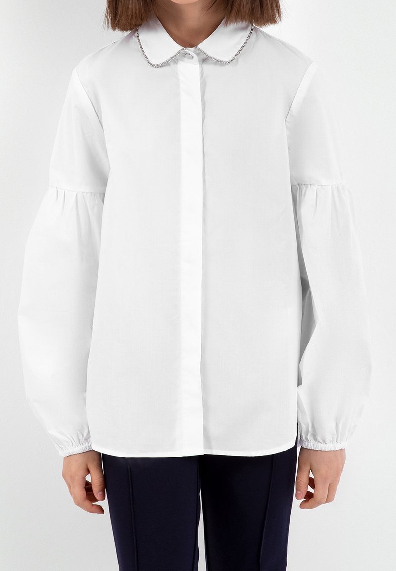 Enfant portant un chemisier blanc à manches longues bouffantes et un pantalon noir, visible des épaules aux hanches sur un fond uni.