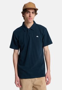Mørkeblå polo-shirt med krave, korte ærmer og et diskret hvidt logo på brystet. Parret med khaki corduroy-bukser.