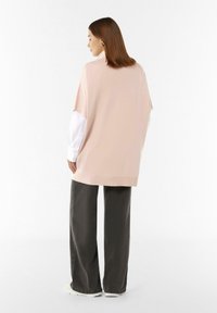 Überdimensioniertes pinkes Strickoberteil mit weitem Ärmel, kombiniert mit einem weißen Hemd, dazu dunkle graue weite Hosen und weiße Sneakers.