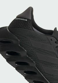 Black athletic shoe with a textured fabric upper, padded collar, and distinctive molded sole featuring oval cutouts and logo detail.