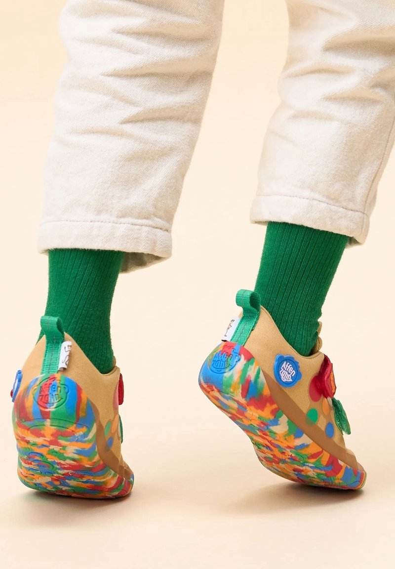 Personne portant des chaussettes vertes et un pantalon beige avec des chaussures slip-on colorées au motif tie-dye, debout sur un fond clair.