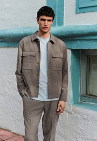 Chaqueta camisa de lino caqui con dos bolsillos en el pecho, botones de latón, combinada con una camiseta azul claro de cuello redondo y pantalones a juego.