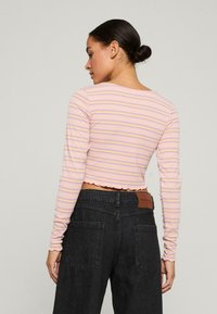 Langärmliges, kurzes Oberteil in hellem Rosa mit dünnen, horizontalen Streifen in Lila. Hat einen gewellten Saum und ein geripptes Muster. Kombiniert mit dunklen Jeans.