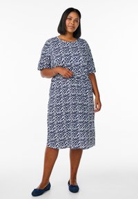 Navy- und weißes gemustertes Kleid mit kurzen Ärmeln, tailliertem Bund, knielanger Länge und rundem Ausschnitt. Kombiniert mit blauen Ballerinas.