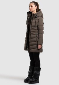 Abrigo acolchado hasta la rodilla en verde oliva con capucha, costuras verticales y bolsillos con cremallera. Combinado con botas de nieve negras forradas de piel.