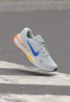 Chaussure de sport blanche avec le logo Nike bleu et des accents orange-jaune, flottant au-dessus d'un passage piéton avec de petits débris en dessous.
