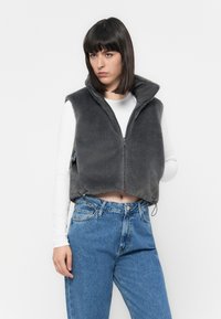 Gilet en fausse fourrure grise avec un col montant, design court, fermeture éclair et attaches latérales, associé à un jean bleu taille haute et un haut blanc à manches longues.