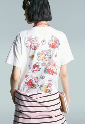 Personne portant un t-shirt blanc avec des personnages de dessins animés colorés et des desserts dans le dos, tenant des cahiers, avec un pull rayé rose noué autour de la taille.