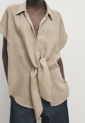 Blouse beige en lin à manches courtes avec un large col et un nœud à l'avant noué à la taille, portée avec un jean denim foncé.