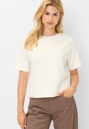 Femme blonde portant un haut blanc à manches courtes et un pantalon marron, une main dans la poche, debout devant un fond blanc uni.