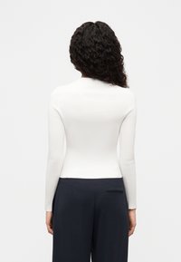 Haut blanc à manches longues côtelé avec une coupe ajustée, porté avec un pantalon sombre. Texture simple et lisse ; aucun motif ou accessoire visible.