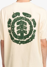 Cremefarvet bomulds T-shirt med et stort grønt trægrafik på ryggen, omkranset af grønne bladdetaljer. Korte ærmer og afslappet pasform.