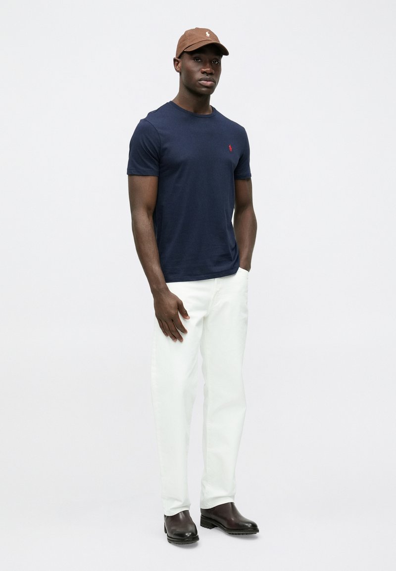 Námornícke tričko s logom, biele nohavice a hnedá baseballová čiapka. Pevné hnedé topánky. Hladká látka, pohodlný strih, jednoduchý dizajn.