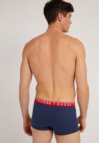 Himbeerfarbene Boxershorts aus Navy-Blau mit rotem GUESS-Logo-Bund, die einen engen Schnitt und eine glatte Textur aufweisen, von hinten sichtbar.