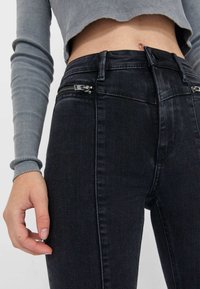 Högmidjade svarta denimjeans med två framfickor med dragkedja och subtila blekningsdetaljer. Tyget är slätt med en åtsittande silhuett.