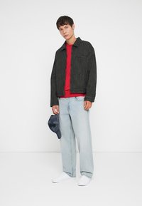 Casaco riscado em preto e branco, camisa vermelha por baixo, jeans folgados em azul claro, ténis brancos, segurando um chapéu azul escuro. Vestuário simples e casual.