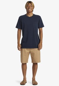 Mörkblå bomullströja med korta ärmar och bröstficka på vänster sida, tillsammans med khakifärgade shorts och bruna flip-flops. Enkel avslappnad outfit.