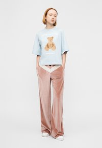 Ανοιχτό μπλε βαμβακερό μπλουζάκι με σχέδιο αρκούδας και κείμενο· συνδυασμένο με ροζ βελούδινο παντελόνι φαρδιάς γραμμής με λευκή λεπτομέρεια σε σχήμα ζιγκ-ζαγκ και λογότυπο.