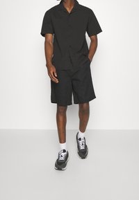 Černá košile s krátkým rukávem a zapínáním na knoflíky, s límečkem, v kombinaci s černými šortkami. Obuv zahrnuje černé tenisky s bílými akcenty a ponožky.