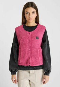 Fleece vest in felroze met een ronde hals, knopen aan de voorkant en een klein logo accent. Draagt over een zwart shirt met lange mouwen.