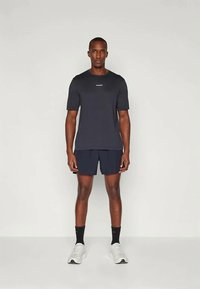 Mies seisoo yksinkertaista taustaa vasten, pukeutuneena mustaan t-paitaan, mustiin shortseihin, mustiin sukkiin ja valkoisiin urheilukenkään, kasvot eteenpäin.