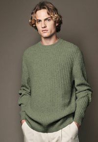 Pull en tricot vert olive avec côtes verticales, col rond et manches longues, associé à un pantalon de couleur claire. Texture douce et coupe décontractée.