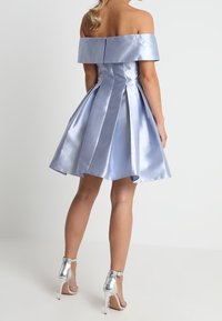 Žena v bezramenných, světle modrých saténových šatech po kolena s plisováním, doplněná stříbrnými lodičkami na vysokém podpatku s páskem kolem kotníku.