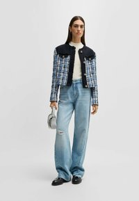 Tweedjakke med navy, blå og hvidt mønster; slidte kanter; runde knapper; parret med lyseblå vide jeans og en lille grå håndtaske.