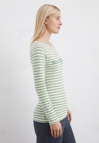 Camicia a maniche lunghe a righe verdi e bianche con scollatura tonda e testo ricamato sul davanti, indossata con pantaloni scuri.