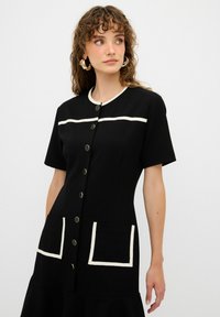 Vestido negro con mangas cortas, cuello redondo, ribete blanco, botones de latón y dos bolsillos frontales con detalles blancos. Textura suave.