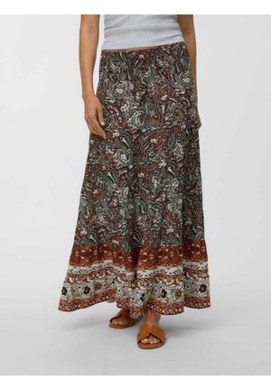 Femme portant une longue jupe à motifs avec des dessins paisley rouges, blancs et noirs et des sandales marron, debout sur un fond uni.