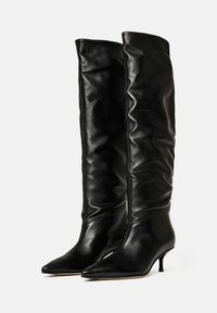 Botas negras de cuero hasta la rodilla con un diseño holgado, punta afilada y tacón kitten bajo, con textura suave y detalles de costura.