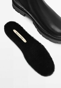 Stivaletto in pelle nera con suola spessa, accompagnato da un sottopiede in peluche nero con un logo su un'etichetta crema.