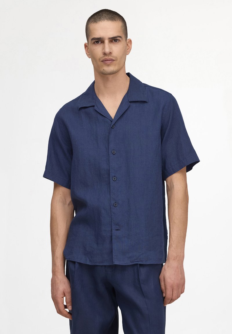 Homme aux cheveux courts portant une chemise bleu foncé à manches courtes boutonnée et un pantalon bleu foncé assorti, debout devant un fond uni.