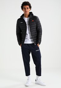 Schwarze Daunenjacke mit Kapuze und vertikalen Nähten, kombiniert mit einem weißen T-Shirt und marineblauen Sporthosen mit weißen Streifen. Weiße Sneakers.