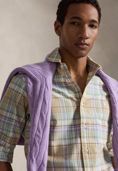 Jeune homme portant une chemise à carreaux pastel boutonnée avec un pull en maille torsadée lavande posé sur ses épaules, sur un fond neutre.
