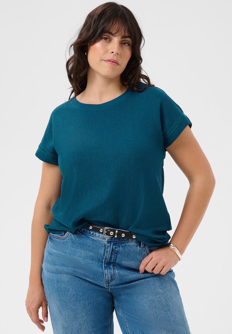 Femme aux cheveux foncés mi-longs portant un haut texturé à manches courtes couleur sarcelle, rentré dans un jean bleu clair avec une ceinture noire.