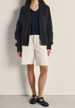 Vrouw draagt een zwarte pluche cardigan, een zwart V-hals topje, knielange witte shorts en zwarte sandalen met gesloten neus, staand tegen een effen muur.