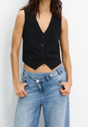 Personne portant un gilet noir sans manches boutonné et un jean bleu clair avec des ceintures qui se chevauchent, mains dans les poches devant un fond uni.
