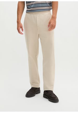 Mann trägt beige gerade geschnittene Hose, schwarze Loafers und ein teilweise sichtbares gestreiftes Hemd auf weißem Hintergrund.