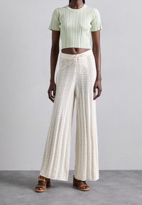 Kobieta w kremowych, szerokich dzianinowych spodniach w kwiatowe wzory, jasnoszarej dzianinowej, krótkiej bluzce oraz brązowych sandałach na obcasie.