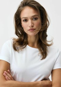 Biały bawełniany T-shirt z krótkimi rękawami, z klasycznym okrągłym dekoltem. Modelka ma średniej długości, falujące brązowe włosy.