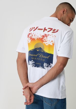 Man draagt een wit T-shirt met een kleurrijke afbeelding van Mount Fuji, Japanse tekst en "Grand Resort Mount Fuji Japan" op de achterkant, handen gekruist achter de rug.