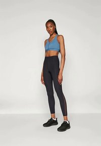 Helesinine spordirinnahoidja, kõrge vöökohaga mustad retuusid, millel on võrkpaneelid, ja mustad spordijalatsid. Modell seisab neutraalse tausta ees.