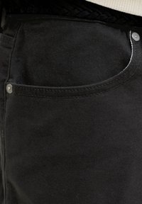 Fekete farmernadrág zsebének közeli képe, látható varrással és fém szegecsekkel a felső sarkokban.