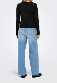 Femme debout, vêtue d’un pull noir avec des sequins sur les épaules, d’un jean large bleu clair et des bottes noires, de dos sur fond blanc.