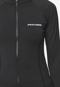 Fekete, hosszú ujjú dzseki elülső cipzárral, varrás nélküli kialakítással és fehér "PEGADOR" logóval a bal oldalon. Rugalmas anyagból készült.